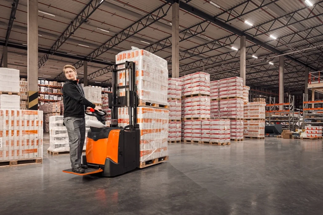 BT Staxio SPE160L, electric stacker with elevating support arms, platform and side guards. Application photo in warehouse environment.